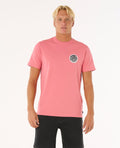 Modelo con camiseta Rip Curl Wettie Passage Icon Tee rosa, vista frontal, estampado pequeño en el pecho, corte estándar.