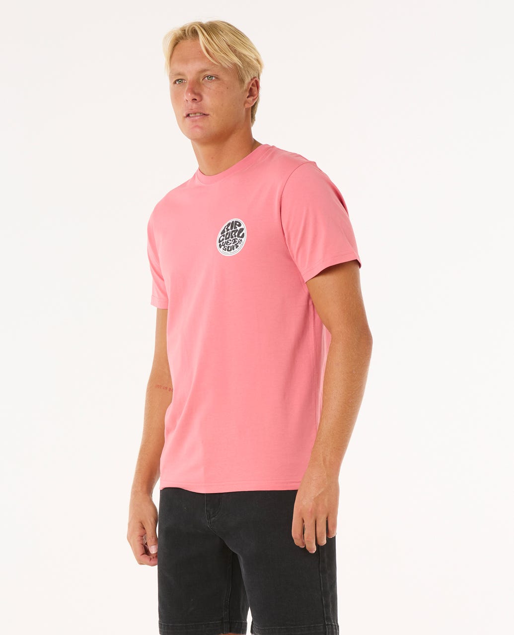 Modelo con camiseta Rip Curl Wettie Passage Icon Tee rosa, vista lateral, ajuste estándar, algodón orgánico.