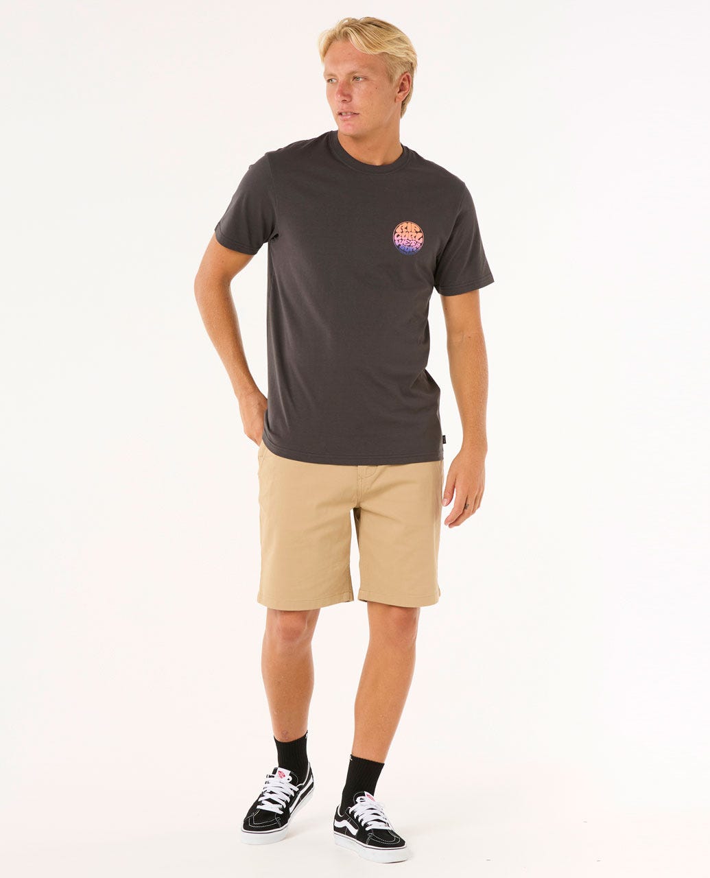 Modelo con camiseta Rip Curl Wettie Passage Icon Tee negra y bermudas beige, look casual surfero.