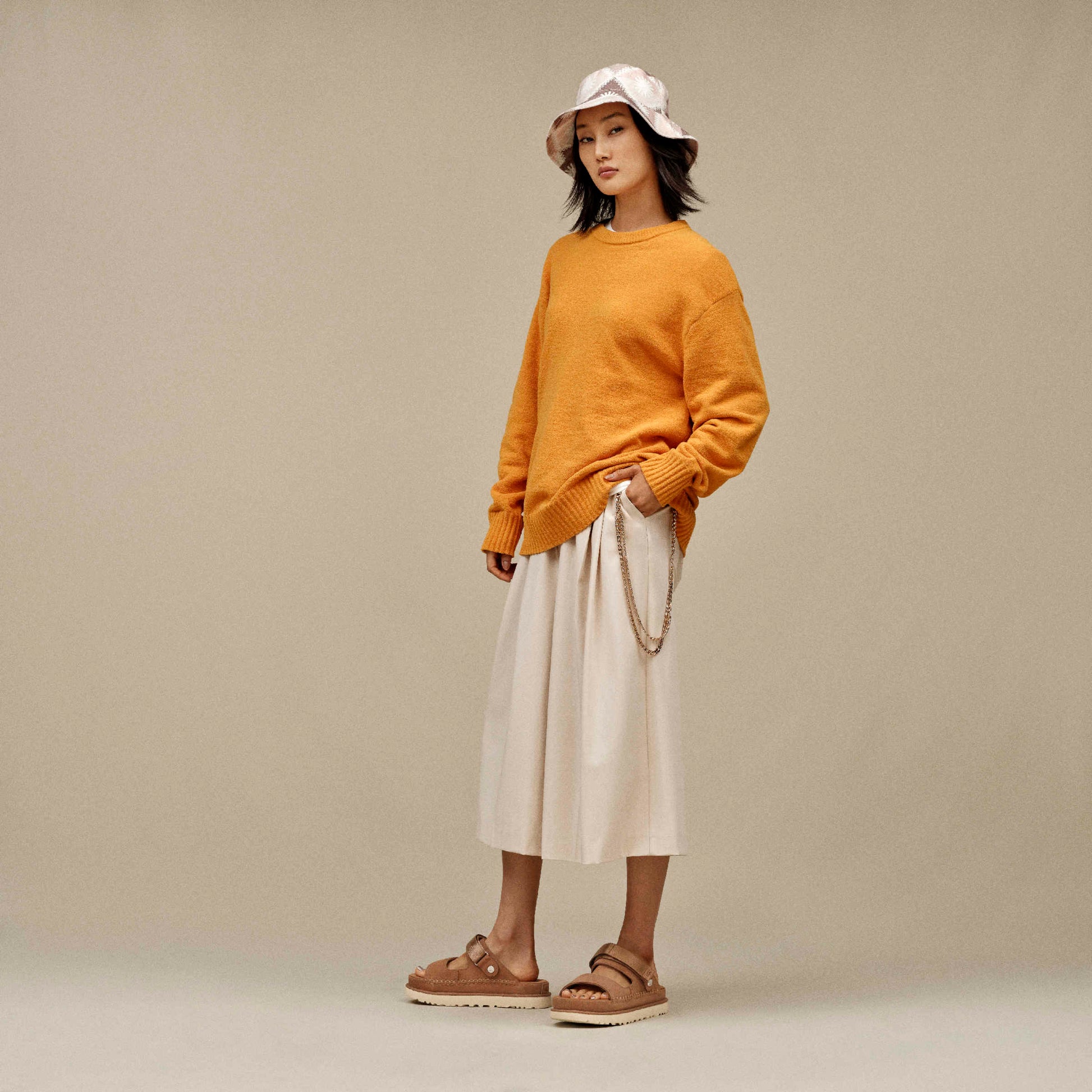 Mujer con look urbano y sandalias UGG Goldenstar Glide marrón oscuro, combinación con falda beige y jersey mostaza