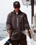 Hombre vistiendo la chaqueta Salty Crew Long Range Jacket negra mientras sostiene peces voladores, perfecta para condiciones marítimas.