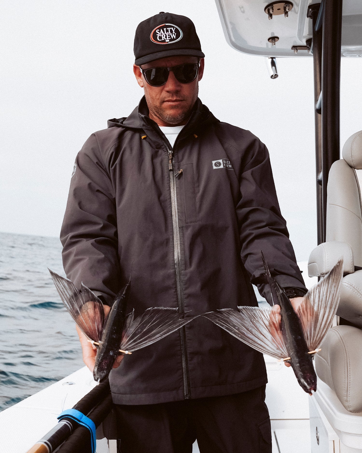 Hombre vistiendo la chaqueta Salty Crew Long Range Jacket negra mientras sostiene peces voladores, perfecta para condiciones marítimas.