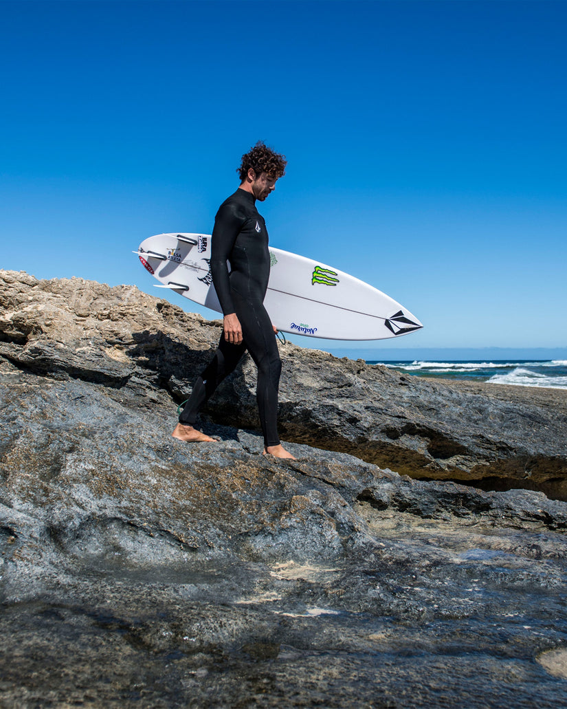 Modelo vistiendo el traje de neopreno VOLCOM 4/3MM Chest Zip Fullsuit en color negro, bajando por unas rocas con su tabla para una sesión de suf