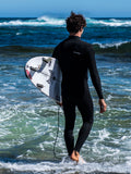 Modelo vistiendo el traje de neopreno VOLCOM 4/3MM Chest Zip Fullsuit en color negro, entrando en el agua con su tabla para una sesión de suf