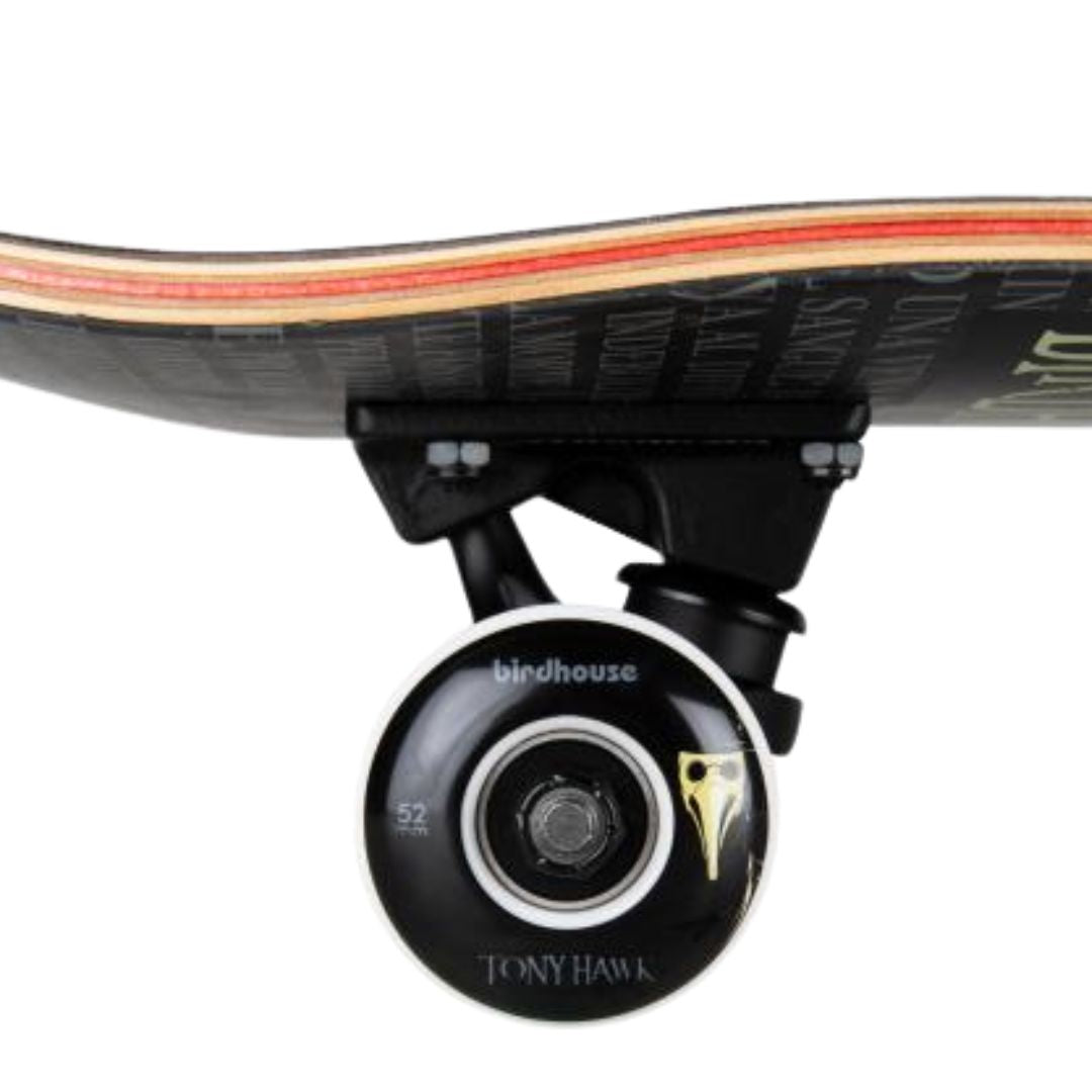 Detalle lateral de una rueda negra de 52 mm con borde blanco, logo Birdhouse, máscara dorada y texto Tony Hawk, truck negro y capas de arce del skate completo Birdhouse Stage 3 Hawk Plague Doctor