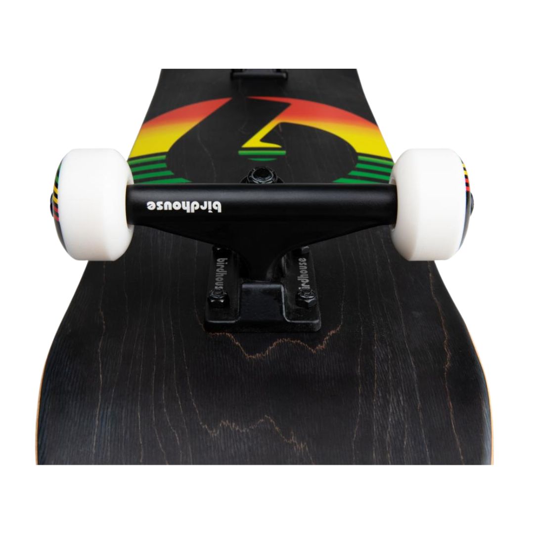 Skate completo Birdhouse Stage 3 Sunset visto desde abajo en perspectiva: truck negro de 5", ruedas blancas, tabla negra con veta de madera y logo B con degradado rojo, amarillo y verde