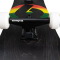 Truck negro con logo Birdhouse y ruedas blancas sobre la tabla negra con veta de madera del skate completo Birdhouse Stage 3 Sunset, con el sol en degradado rojo, amarillo y verde