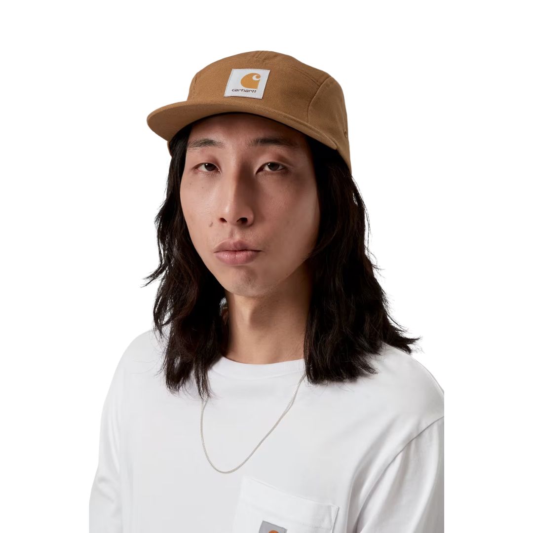Carhartt WIP Backley Cap en color hamilton brown sobre modelo masculino, vista frontal. Gorra de cinco paneles en lona de algodón marrón caramelo con visera plana, etiqueta cuadrada tejida Carhartt WIP en el panel frontal y ajuste regular. Combinada con camiseta blanca de manga larga con bolsillo en el pecho.