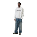 Modelo de cuerpo entero con la sudadera Carhartt WIP Carhartt Sweat en color Ash Heather / Graphite, combinada con vaqueros anchos desgastados y zapatillas.