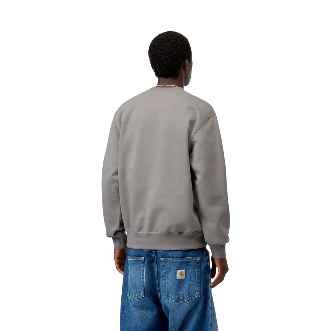 Vista trasera de modelo luciendo la sudadera Carhartt WIP Carhartt Sweat en Shady Grey, mostrando el ajuste oversize en hombros y mangas junto a vaqueros Carhartt WIP con parche trasero.