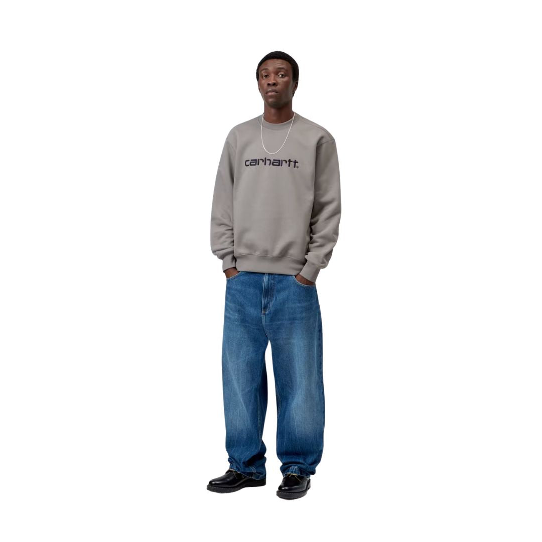 Modelo con sudadera Carhartt WIP Carhartt Sweat en gris Shady Grey y logo Dark Navy combinada con vaqueros anchos, mostrando el calce oversize y la caída relajada de la prenda.