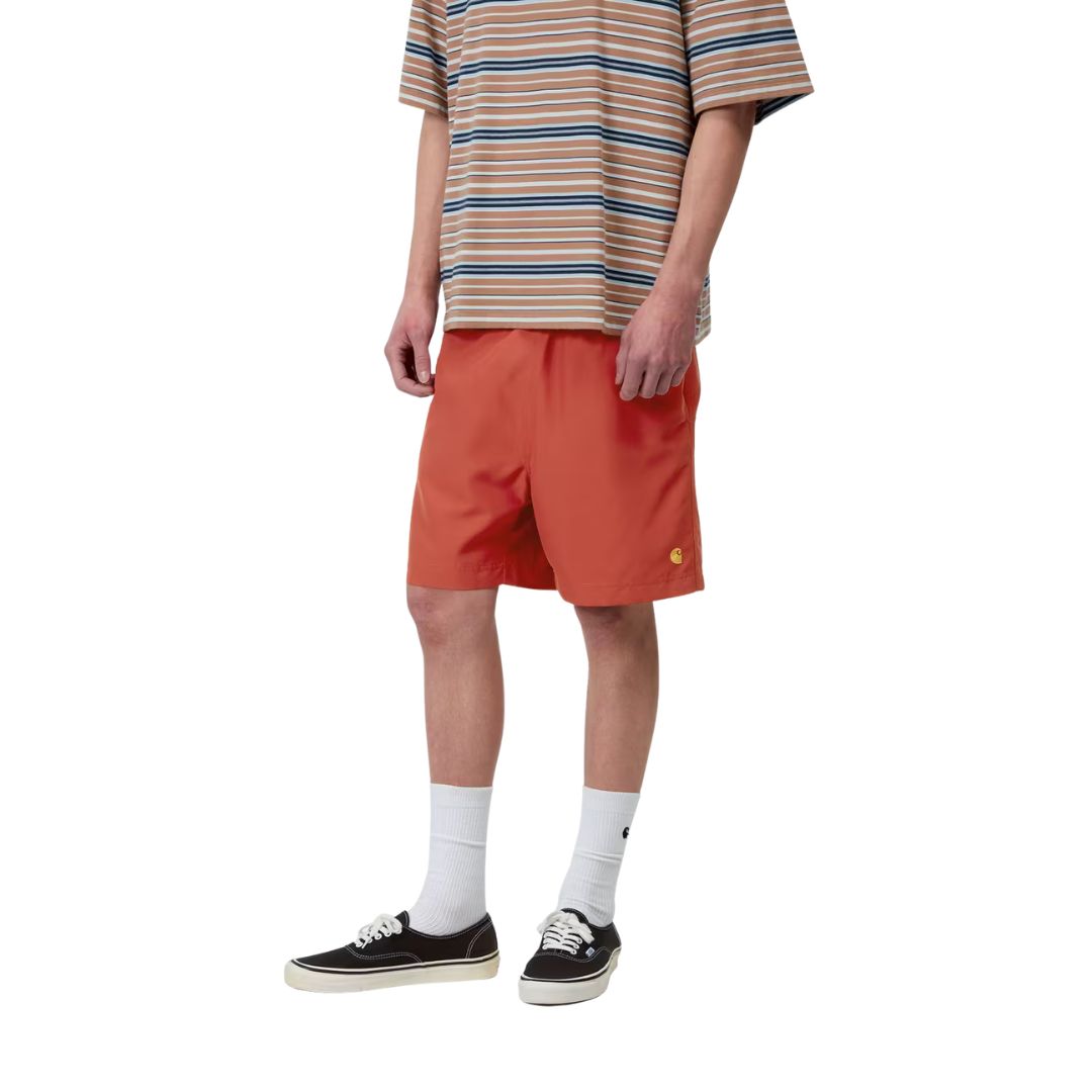 Carhartt WIP Chase Swim Trunks en color oxide red con detalle dorado, vista frontal lateral sobre modelo masculino. Bañador de poliéster ligero en rojo teja con logotipo "C" de Carhartt WIP bordado en dorado en la pierna izquierda, cintura elástica con cordón ajustable y bolsillos laterales integrados.