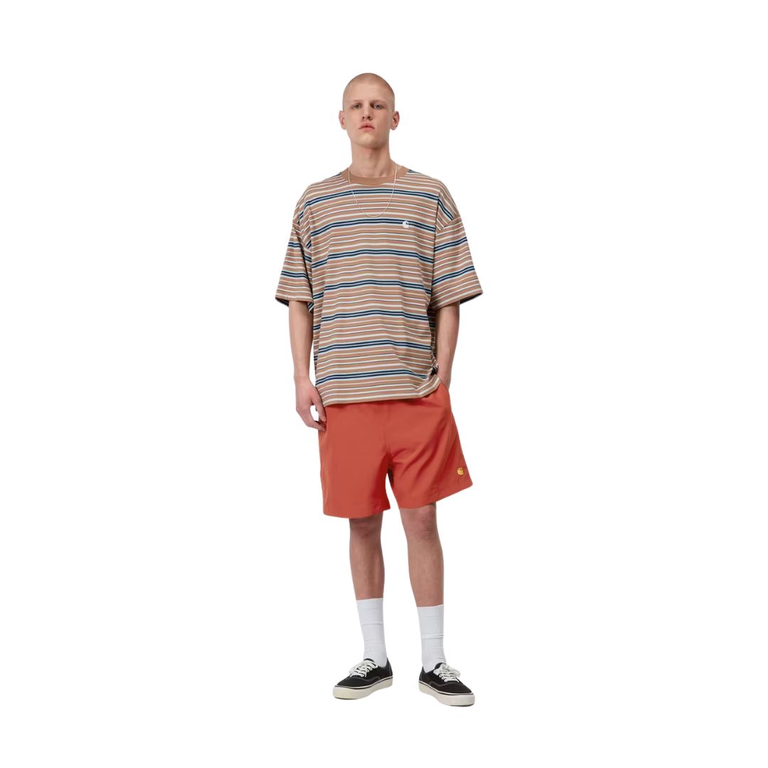 Carhartt WIP Chase Swim Trunks en color oxide red con detalle dorado, vista completa de look sobre modelo masculino. Bañador de secado rápido en rojo teja con "C" bordada en dorado, combinado con camiseta oversize de rayas Carhartt WIP en tonos beige, azul y marrón, calcetines blancos y zapatillas Vans negras.