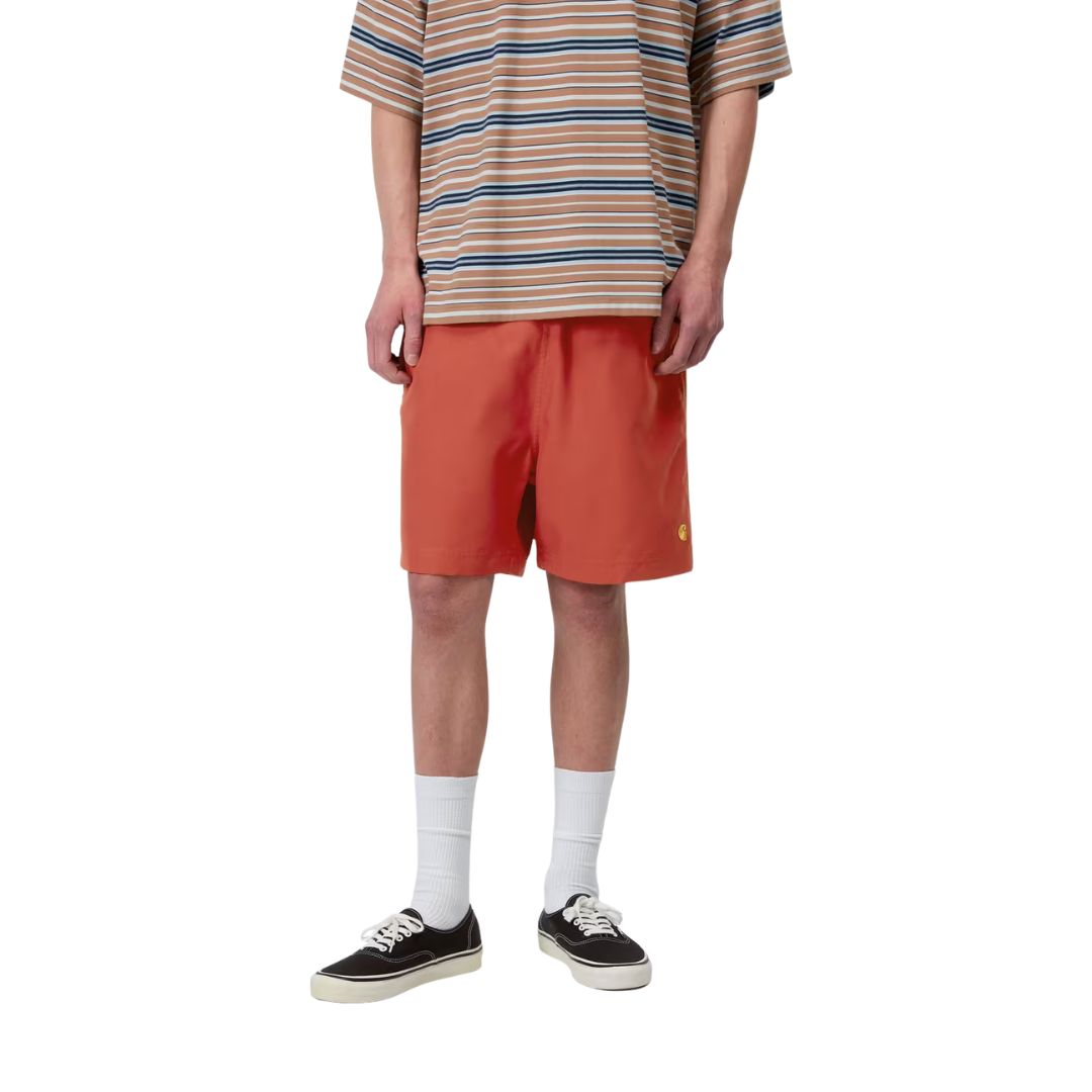 Carhartt WIP Chase Swim Trunks en color oxide red con detalle dorado, vista frontal sobre modelo masculino. Bañador de poliéster repelente al agua en rojo teja con logotipo "C" bordado en dorado en la pierna izquierda y cinturilla elástica con cordón ajustable. Corte regular fit.