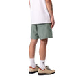 Carhartt WIP Chase Swim Trunks en color velvet green con detalle dorado, vista trasera lateral sobre modelo masculino. Bañador de poliéster ligero en verde apagado con bolsillo trasero con portallaves y cinturilla elástica visible. Corte regular fit.