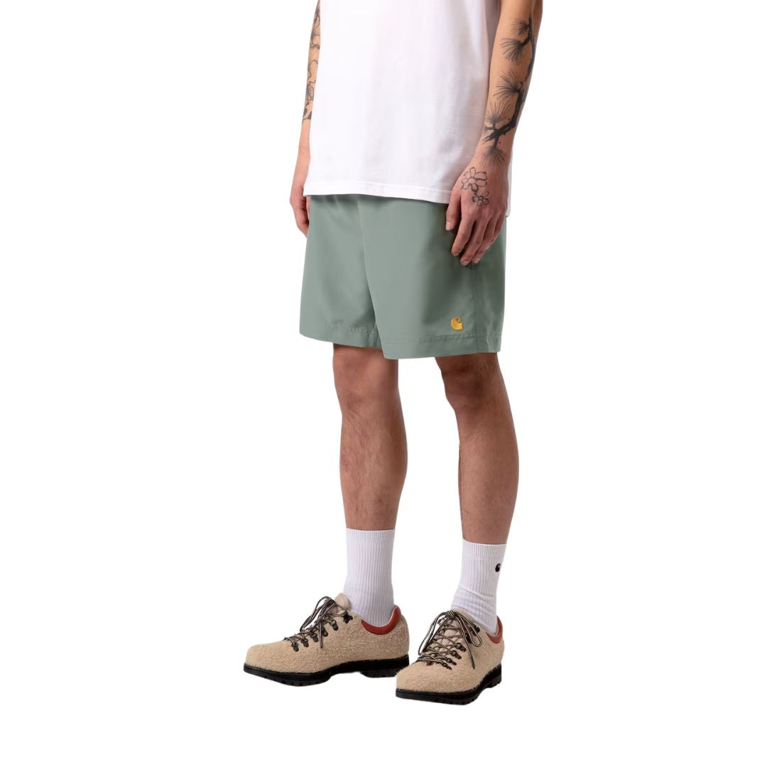 Carhartt WIP Chase Swim Trunks en color velvet green con detalle dorado, vista frontal lateral sobre modelo masculino. Bañador de secado rápido en verde apagado con logotipo "C" de Carhartt WIP bordado en dorado en la pierna izquierda, cintura elástica con cordón y bolsillos laterales integrados.