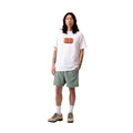 Carhartt WIP Chase Swim Trunks en color velvet green con detalle dorado, vista completa de look sobre modelo masculino. Bañador de poliéster repelente al agua en verde apagado combinado con camiseta blanca con estampado gráfico Carhartt WIP, calcetines blancos y botines de ante beige.
