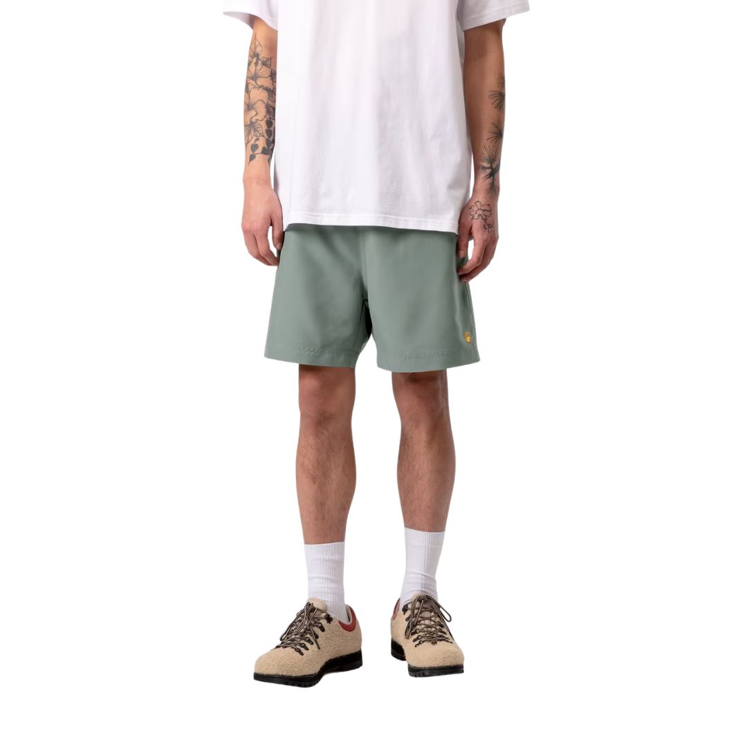 Carhartt WIP Chase Swim Trunks en color velvet green con detalle dorado, vista frontal sobre modelo masculino. Bañador de poliéster ligero en verde apagado con logotipo "C" bordado en dorado en la pierna izquierda y cinturilla elástica con cordón ajustable. Corte regular fit.