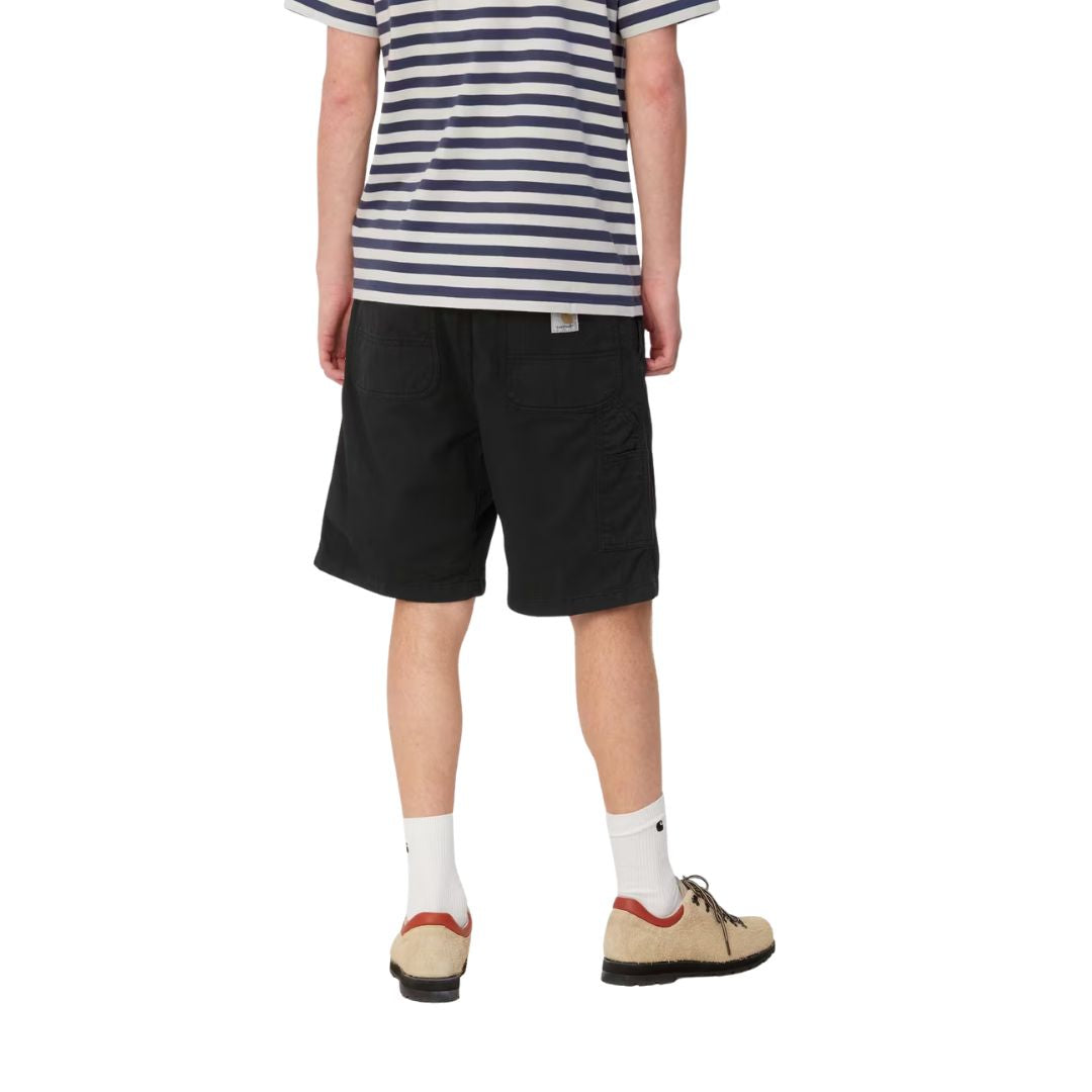 Carhartt WIP Flint Short en color negro teñido en prenda, vista trasera sobre modelo masculino. Bermuda de sarga de algodón orgánico con dos bolsillos traseros de parche con costuras reforzadas, bolsillo para herramientas en la pierna derecha y etiqueta cuadrada tejida Carhartt WIP. Corte regular fit.