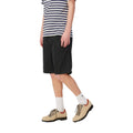 Carhartt WIP Flint Short en color negro teñido en prenda, vista lateral sobre modelo masculino. Bermuda de algodón orgánico con cinturilla elástica, largo por encima de la rodilla y caída limpia de sarga Moraga. Combinada con camiseta de rayas y botines de ante beige.