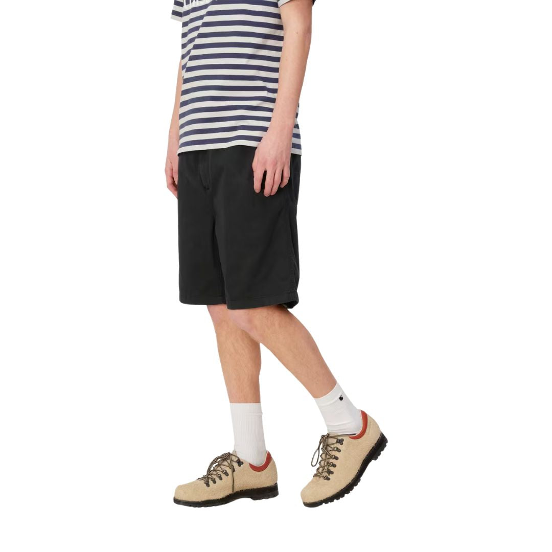 Carhartt WIP Flint Short en color negro teñido en prenda, vista lateral sobre modelo masculino. Bermuda de algodón orgánico con cinturilla elástica, largo por encima de la rodilla y caída limpia de sarga Moraga. Combinada con camiseta de rayas y botines de ante beige.