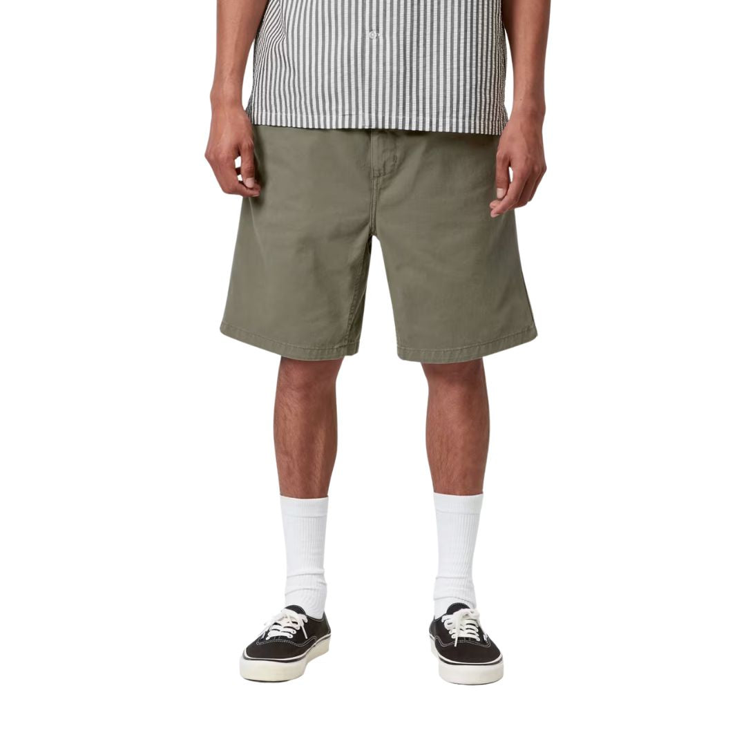 Carhartt WIP Flint Short en color verde leaf teñido en prenda, vista frontal sobre modelo masculino. Bermuda de algodón orgánico con cinturilla elástica con cordón, bolsillos frontales y largo holgado por encima de la rodilla. Corte regular fit combinado con camisa de rayas y zapatillas Vans negras.