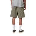 Carhartt WIP Flint Short en color verde leaf teñido en prenda, vista trasera sobre modelo masculino. Bermuda de sarga de algodón orgánico con dos grandes bolsillos traseros de parche, bolsillo para herramientas en la pierna derecha y cinturilla elástica visible. Corte regular fit.