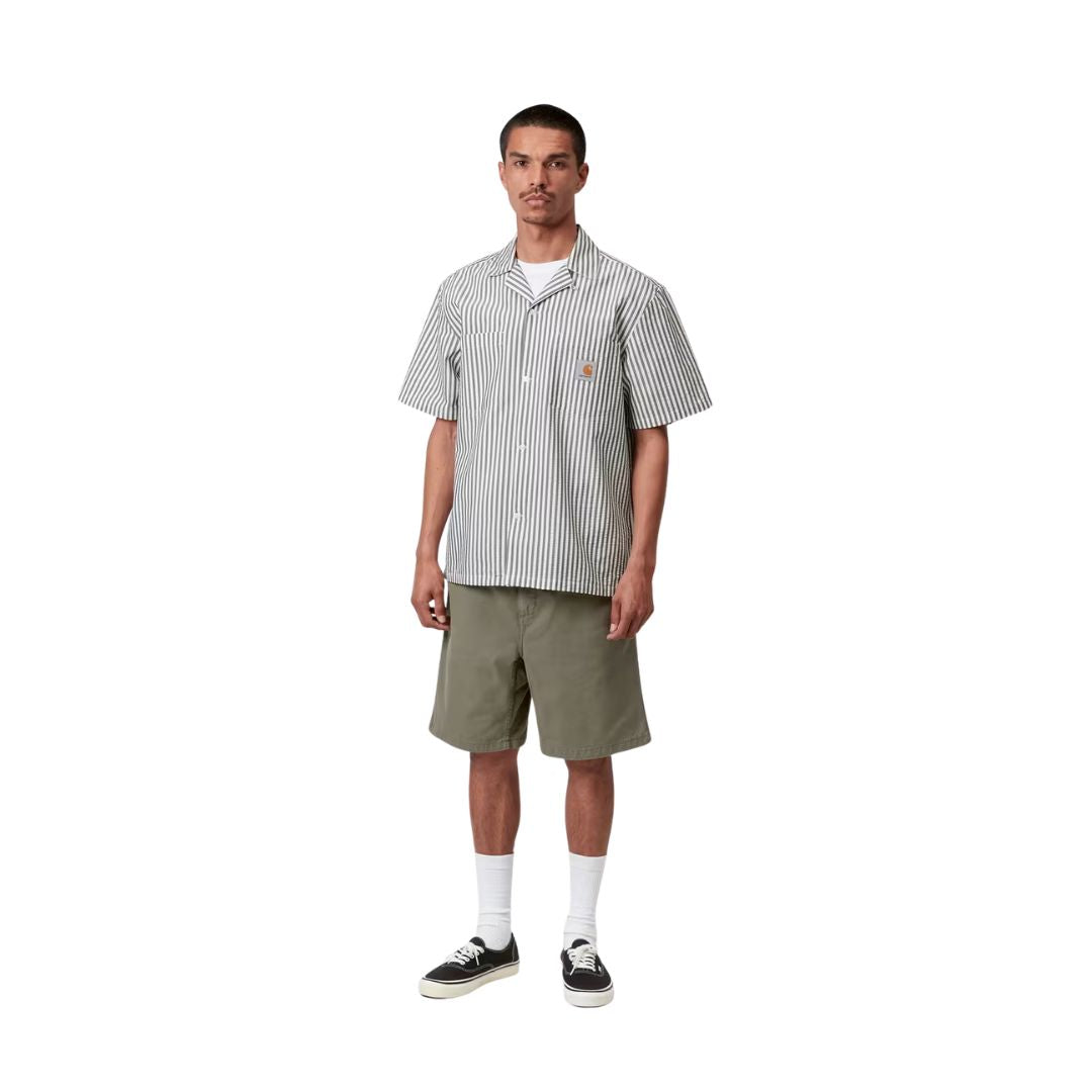 Carhartt WIP Flint Short en color verde leaf teñido en prenda, vista completa de look sobre modelo masculino. Bermuda de sarga orgánica de corte regular combinada con camisa de rayas Carhartt WIP, calcetines blancos y zapatillas Vans negras.