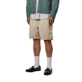Carhartt WIP Flint Short en color wall teñido en prenda, vista frontal lateral sobre modelo masculino. Bermuda de algodón orgánico en tono beige con cinturilla elástica con cordón y largo holgado por encima de la rodilla. Combinada con chaqueta Carhartt WIP verde y camisa de rayas.