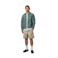 Carhartt WIP Flint Short en color wall teñido en prenda, vista completa de look sobre modelo masculino. Bermuda de sarga orgánica en beige arena combinada con chaqueta Carhartt WIP verde, camisa de rayas, calcetines blancos y mocasines negros de cuero.