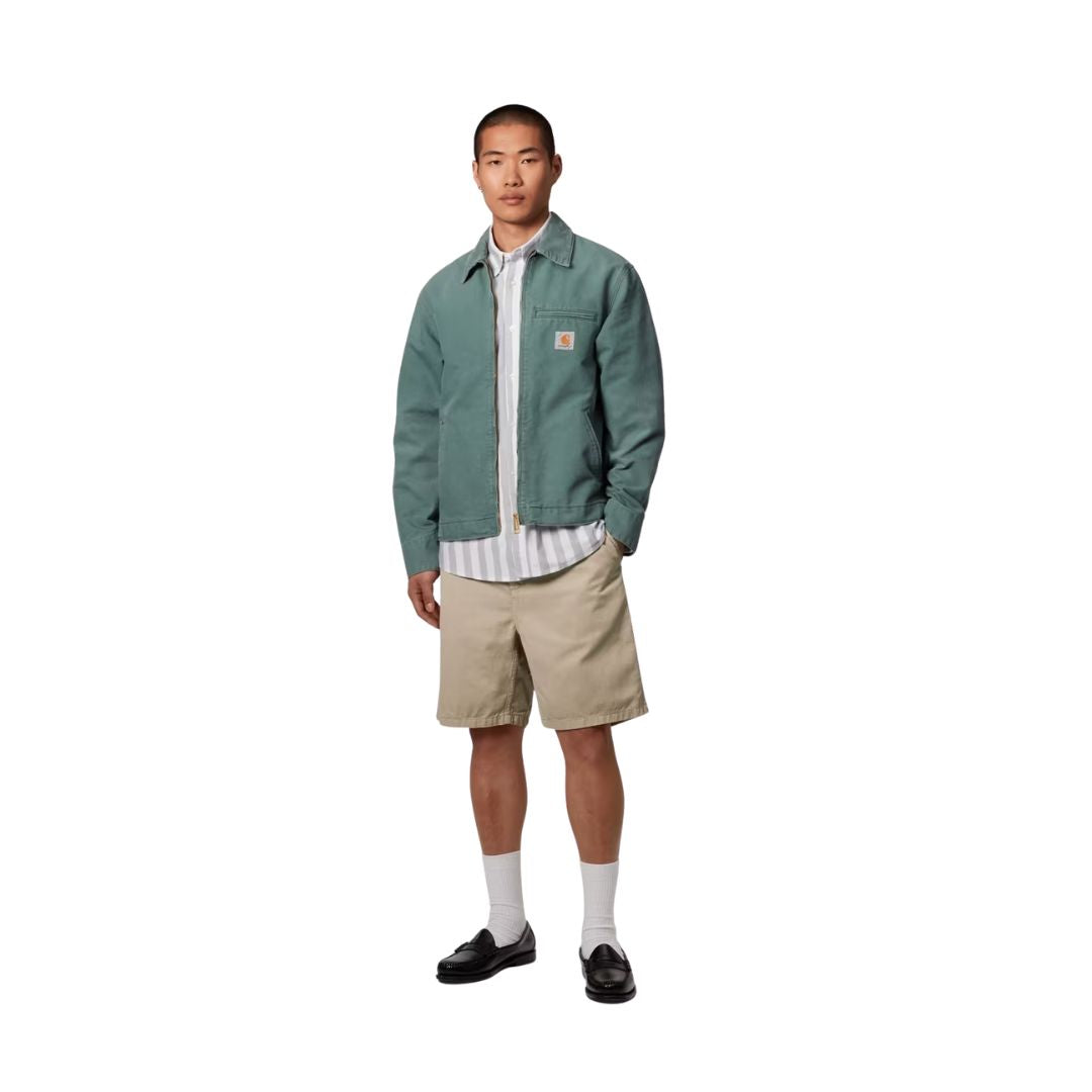 Carhartt WIP Flint Short en color wall teñido en prenda, vista completa de look sobre modelo masculino. Bermuda de sarga orgánica en beige arena combinada con chaqueta Carhartt WIP verde, camisa de rayas, calcetines blancos y mocasines negros de cuero.