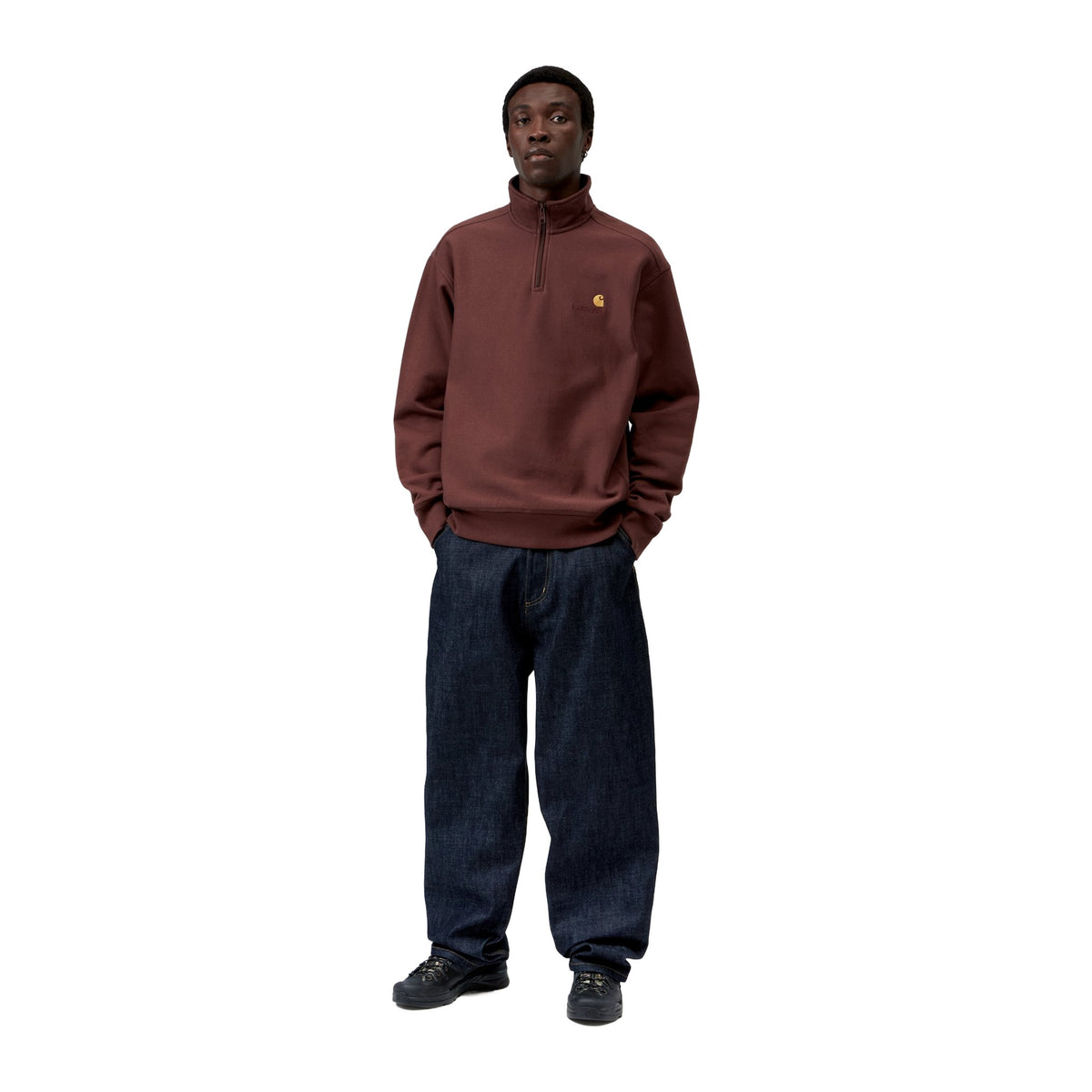 Modelo de cuerpo entero con la sudadera Carhartt WIP Half Zip American Script Sweat en color Amarone, combinada con vaqueros oscuros anchos y zapatillas negras.