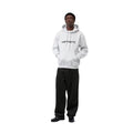 Modelo con la sudadera Carhartt WIP Hooded Carhartt Sweatshirt en Ash Heather y logo Graphite, combinada con pantalón negro ancho y gorro. Corte holgado de estilo urbano.