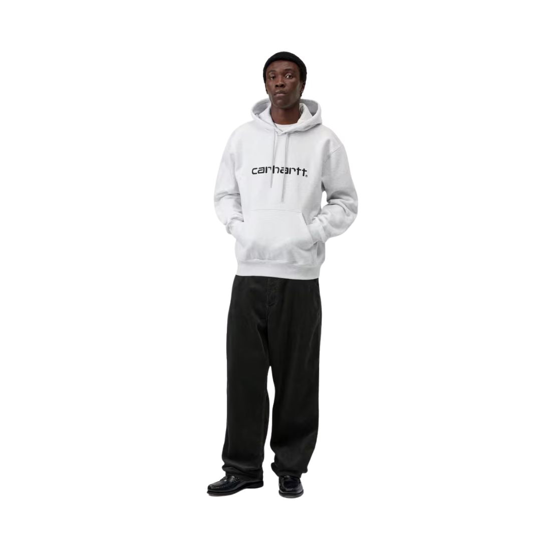 Modelo con la sudadera Carhartt WIP Hooded Carhartt Sweatshirt en Ash Heather y logo Graphite, combinada con pantalón negro ancho y gorro. Corte holgado de estilo urbano.