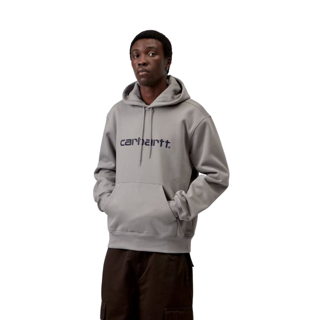 Modelo con sudadera capucha Carhartt WIP Hooded Carhartt Sweat color Shady Grey y logo bordado Dark Navy, cordones ajustables al tono y bolsillo canguro frontal con manos dentro.