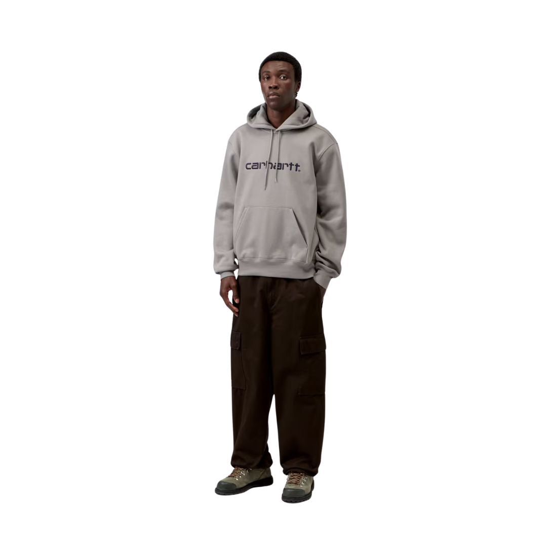 Modelo con sudadera con capucha Carhartt WIP Hooded Carhartt Sweat en Shady Grey combinada con pantalón cargo marrón y botas, mostrando el calce oversize completo de la prenda.