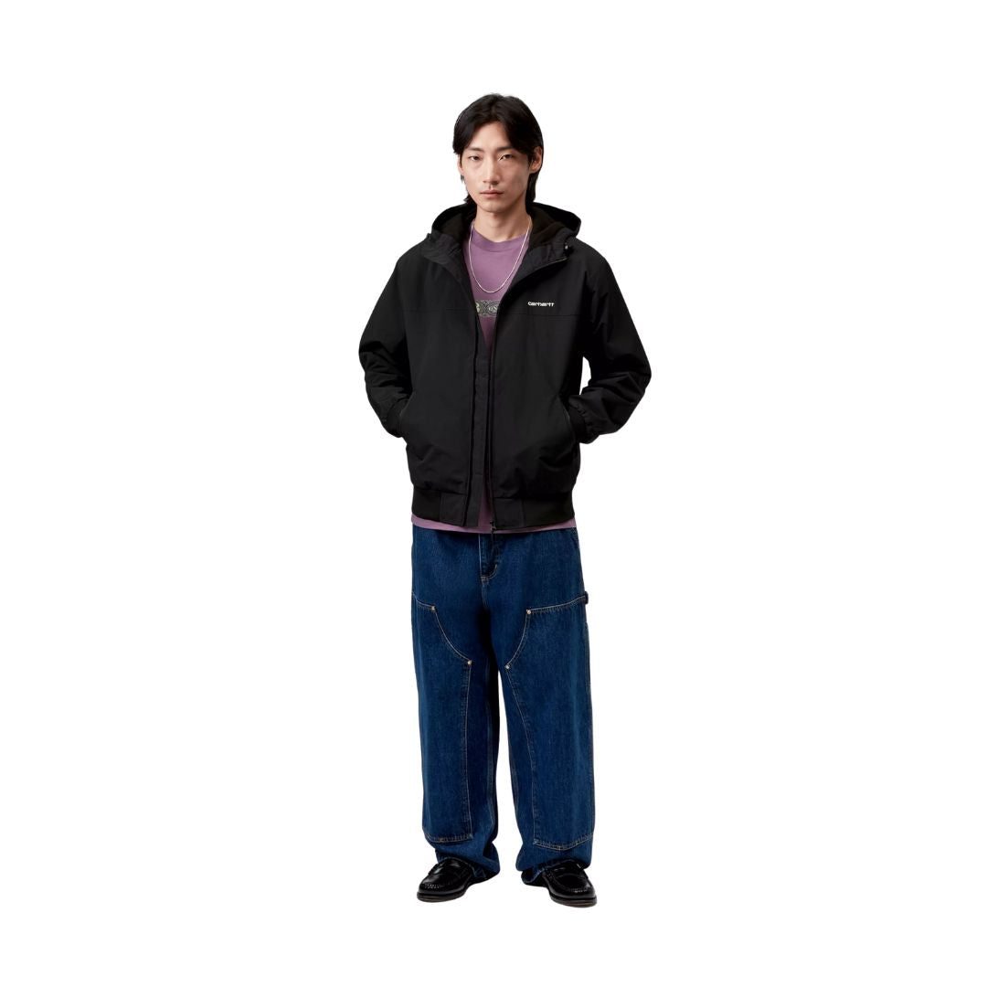 Modelo vistiendo la chaqueta Carhartt WIP Hooded Sail Jacket negra con capucha, combinada con vaqueros anchos y camiseta morada.