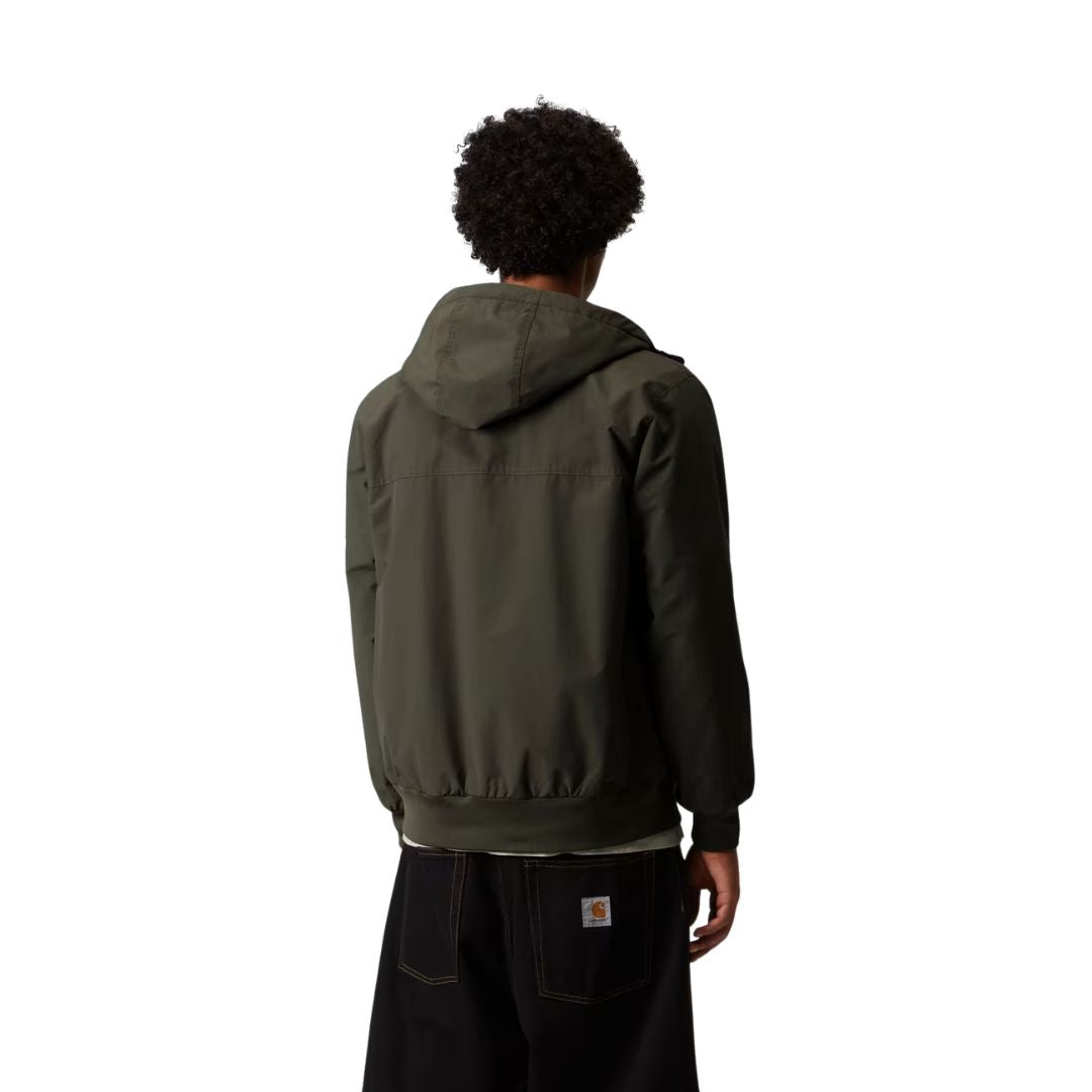 Vista trasera de la chaqueta Hooded Sail Cypress, con capucha, costura de yoke en la espalda y cintura acanalada, sobre pantalón negro con la etiqueta Carhartt WIP en el bolsillo.