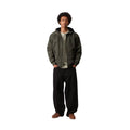 Modelo de pie, de cuerpo entero, con la chaqueta Hooded Sail Cypress abierta sobre camiseta verde claro, pantalón negro muy ancho y zapatos marrones, con las manos en los bolsillos.