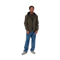 Modelo de pie, de cuerpo entero, con la chaqueta Carhartt WIP Hooded Sail Cypress abierta sobre camiseta negra, vaqueros azules holgados y zapatillas grises.