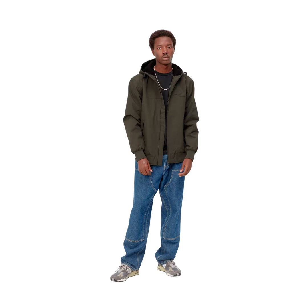 Modelo de pie, de cuerpo entero, con la chaqueta Carhartt WIP Hooded Sail Cypress abierta sobre camiseta negra, vaqueros azules holgados y zapatillas grises.