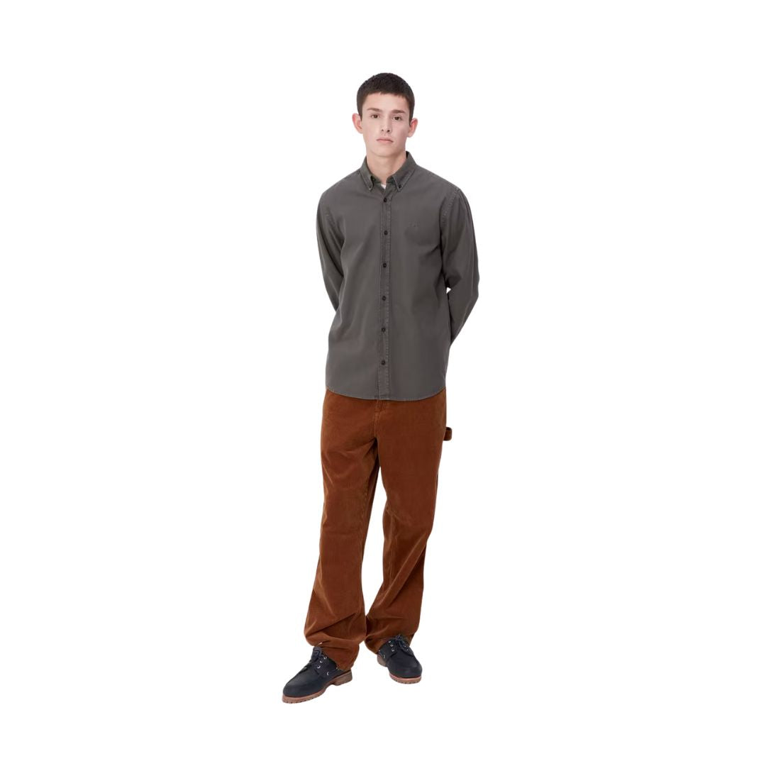 Modelo masculino de pie vistiendo la camisa Carhartt WIP L/S Bolton teñida en prenda color Graphite, combinada con pantalones de pana marrón.