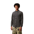 Modelo en plano medio con la camisa de pana Carhartt Madison gris oscuro abrochada, cuello abotonado, puños remangados y pequeño logo bordado en el pecho, con pantalón de camuflaje.