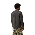 Espalda de un modelo con la camisa de pana Carhartt Madison en gris oscuro, con canesú y pliegue central; se ve la etiqueta naranja de Carhartt en el bolsillo del pantalón cargo de camuflaje.
