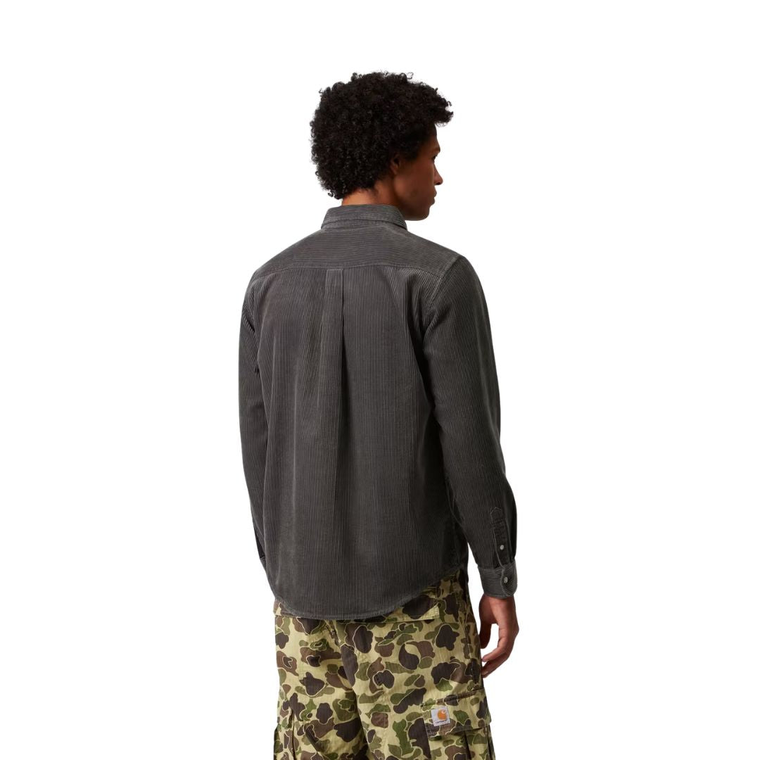 Espalda de un modelo con la camisa de pana Carhartt Madison en gris oscuro, con canesú y pliegue central; se ve la etiqueta naranja de Carhartt en el bolsillo del pantalón cargo de camuflaje.