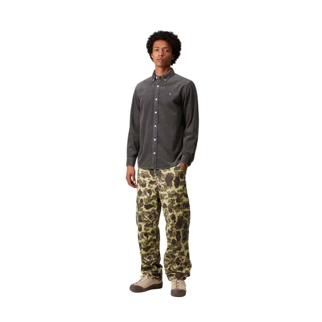 Modelo con la camisa de pana Carhartt Madison gris oscuro, manga larga y cuello abotonado, combinada con pantalón cargo de camuflaje verde y zapatillas de montaña, de pie y en pose relajada.