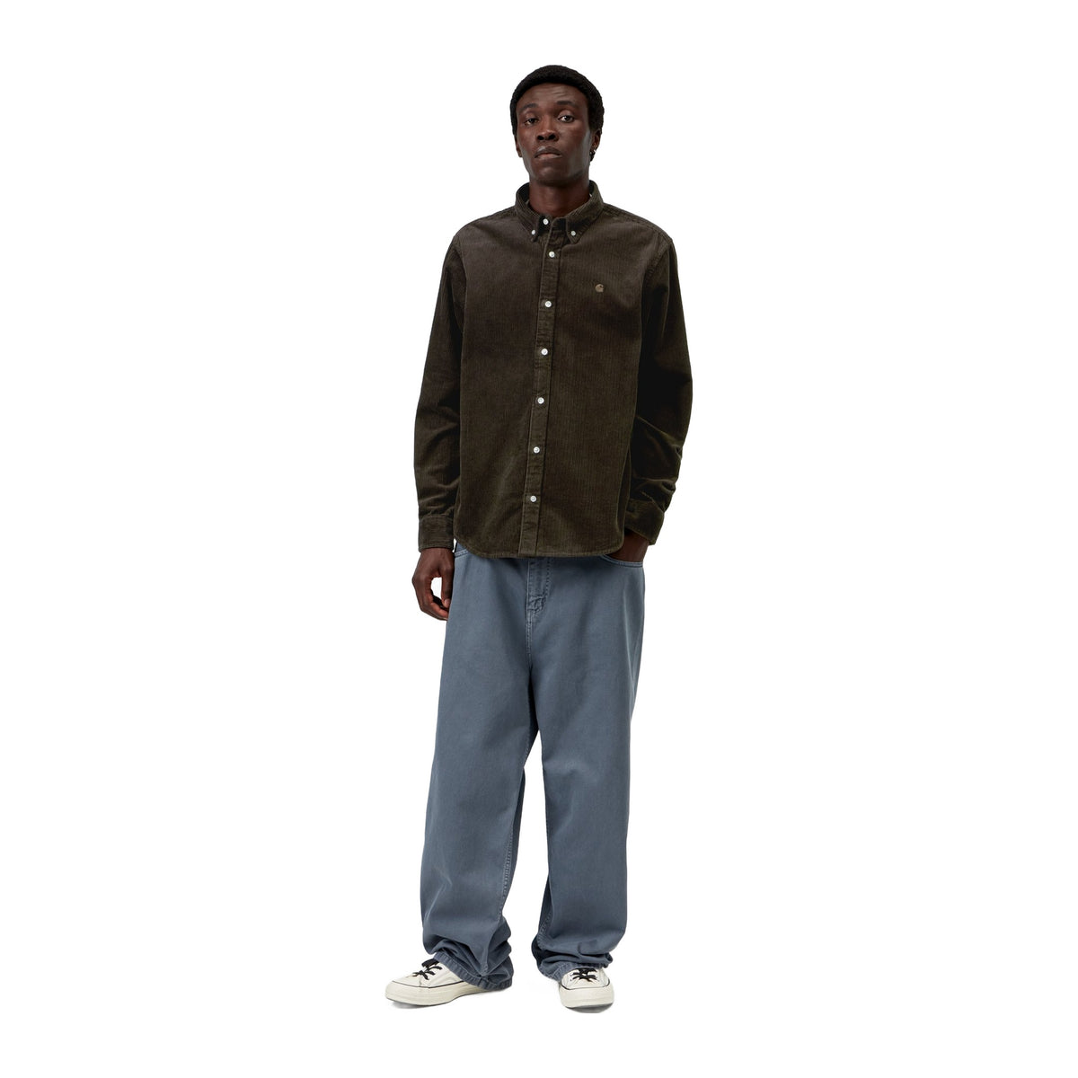 Modelo de cuerpo entero con la camisa de pana Carhartt WIP L/S Madison Cord Shirt en color Tobacco, combinada con pantalón azul grisáceo ancho y zapatillas blancas.