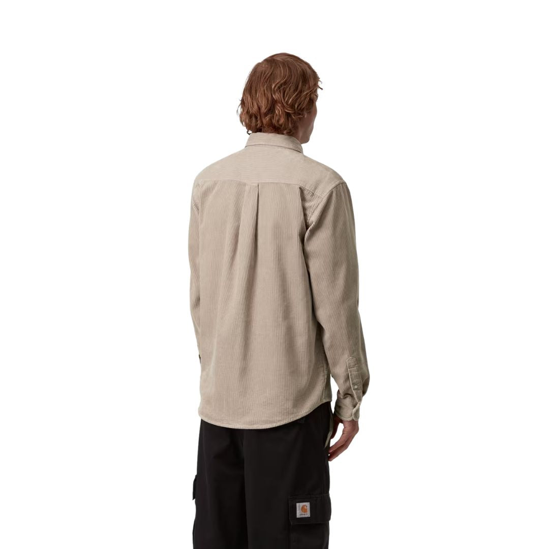 Espalda de un modelo con la camisa de pana Carhartt Madison en beige, con pliegue central y canesú; se ve la etiqueta naranja de Carhartt en el bolsillo del pantalón cargo negro.