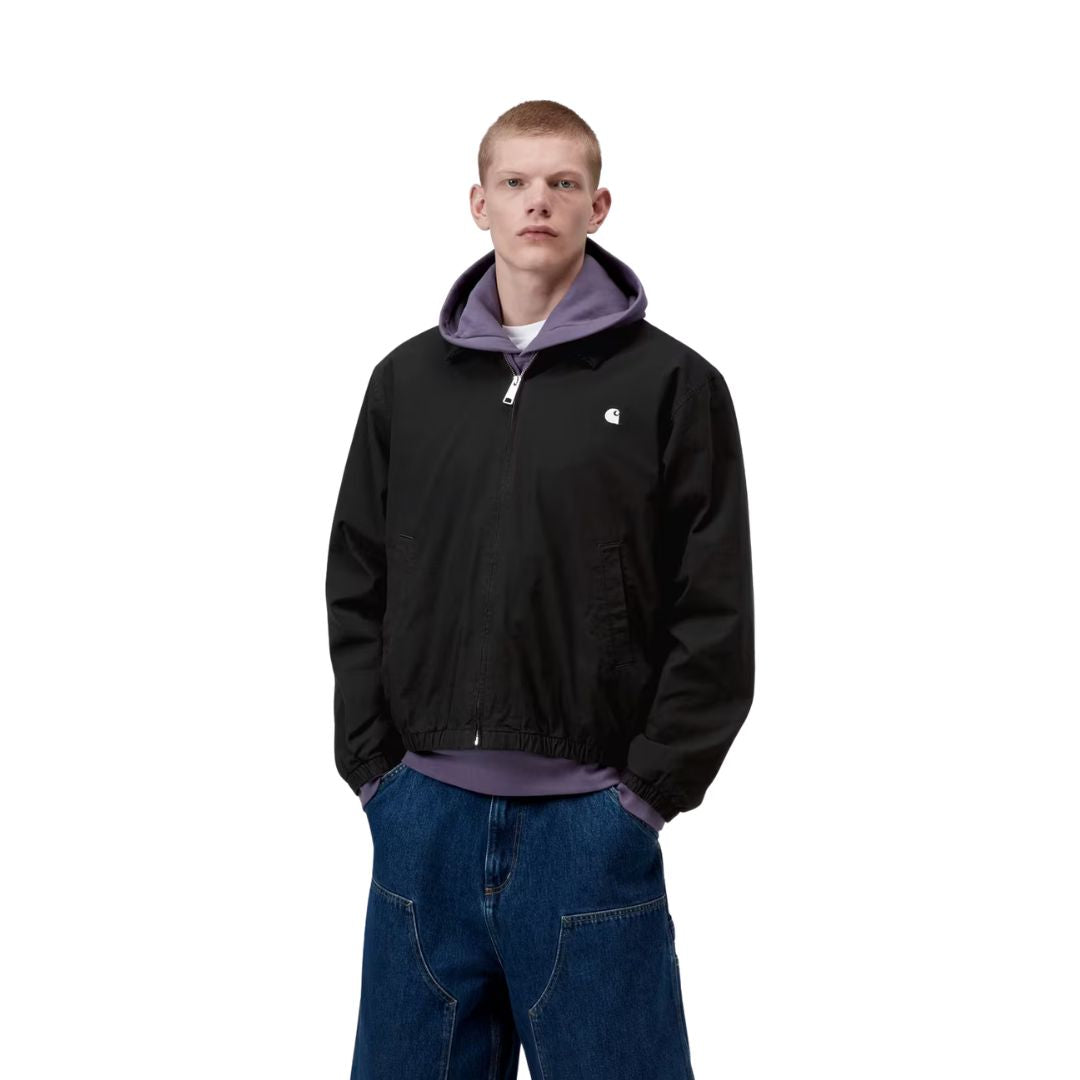 Carhartt WIP Madison Ripstop Jacket negro wax, modelo masculino de frente con cremallera cerrada y capucha lila visible, detalle del bajo elástico y bolsillo lateral.
