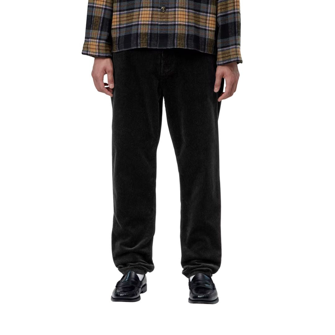 Plano frontal recortado de un modelo con el pantalón de pana Carhartt WIP Newel Pant en color negro, camisa de cuadros y mocasines, resaltando la caída recta del canalé de la pana.